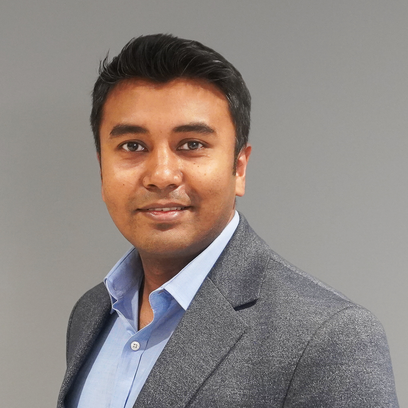 A man with short dark hair wearing a grey blazer and light blue shirt smiles in front of a plain grey background.