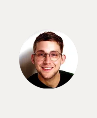 A young man with short brown hair and glasses smiles at the camera. He is wearing a dark shirt and is set against a plain white background.