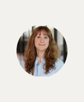 A woman with long, wavy auburn hair wearing a light blue shirt smiles at the camera, standing indoors with a blurred background.