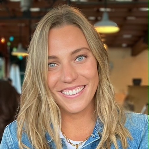 A young woman with long blonde hair and blue eyes smiles at the camera. She is wearing a blue top and a necklace, and is indoors with warm lighting and blurred background details.
