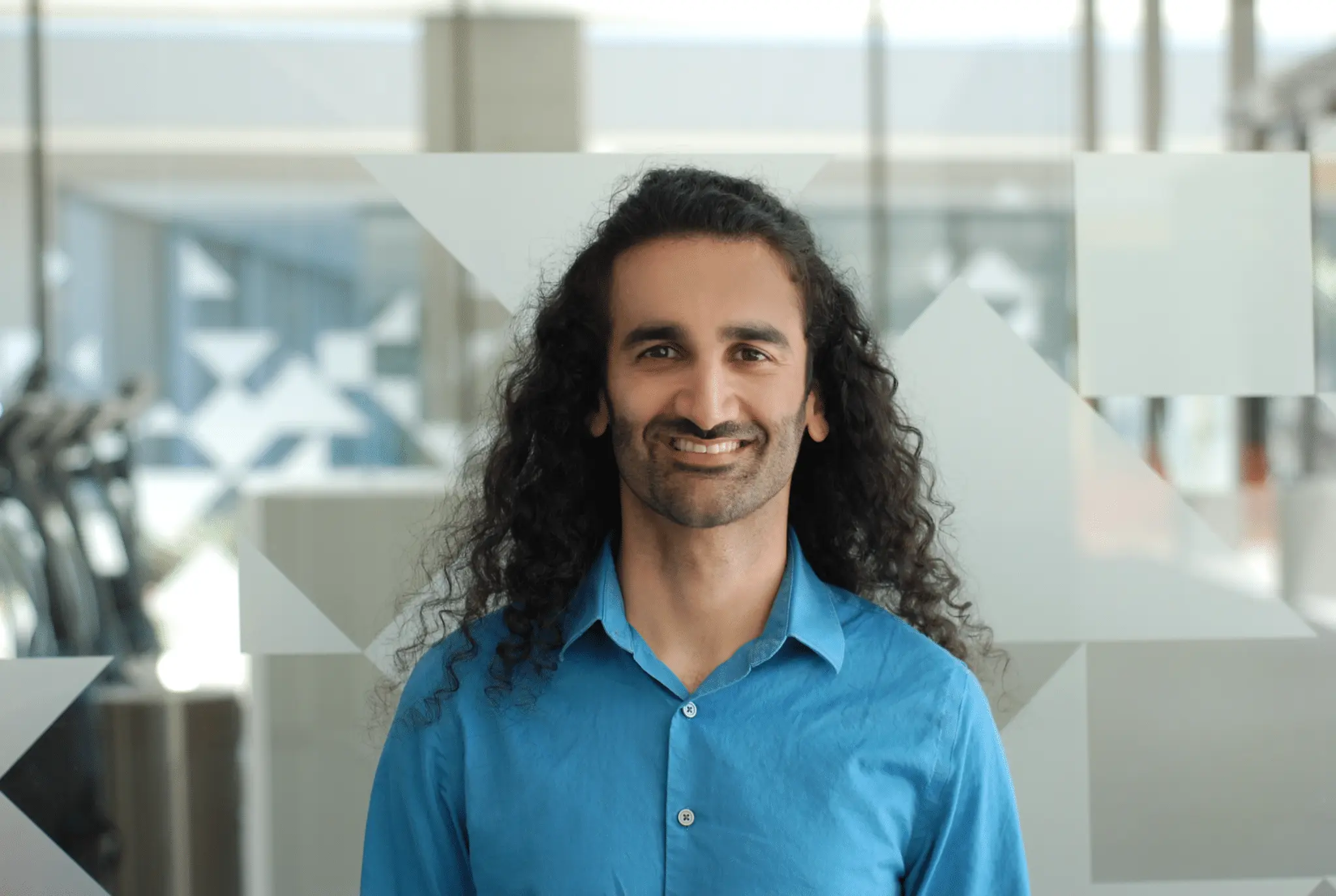 A man with long curly hair and a beard, wearing a bright blue shirt, smiles while standing indoors in front of a patterned glass wall.