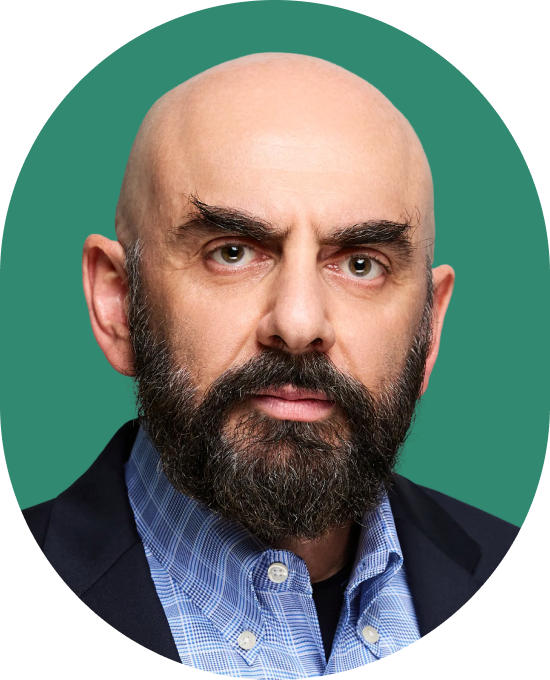 A bald man with a full dark beard wearing a blue checkered shirt and dark blazer, looking directly at the camera with a neutral expression against a green background.