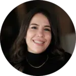 A woman with long dark hair, wearing a black top and a thin necklace, is smiling at the camera against a softly blurred dark background—perfect for showcasing an approachable evisort alternative.