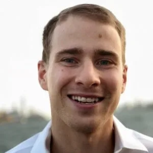A young man with short brown hair and light skin smiles at the camera. He’s wearing a white collared shirt, standing outside with greenery and sky behind him, giving off an outlaw alternative vibe.