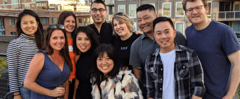 A group of ten adults, including Florence Chan, smiling and posing together outdoors on a rooftop with city buildings in the background. They appear to be enjoying a social gathering on a sunny day.