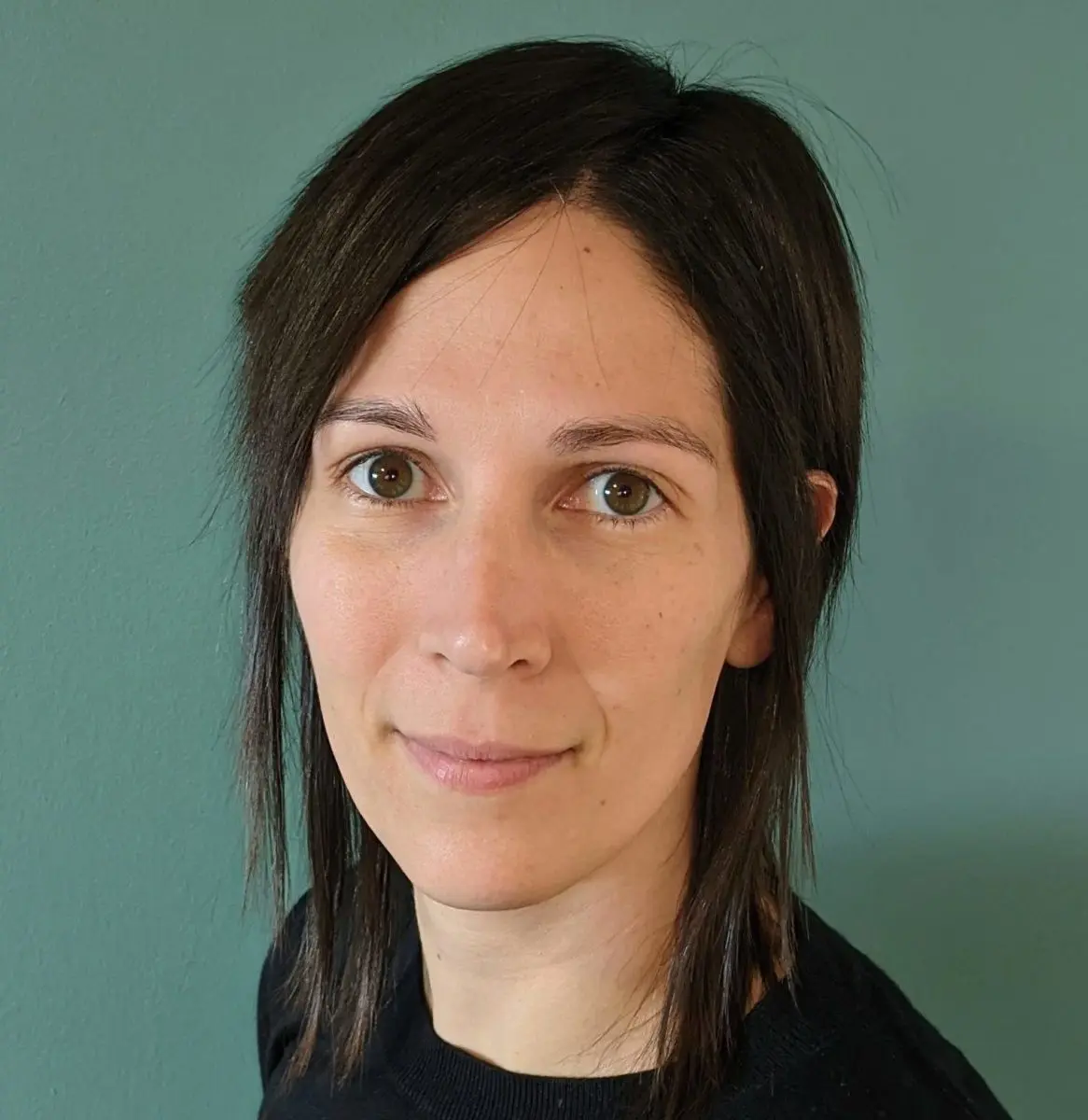 A woman with straight, dark brown hair and brown eyes looks at the camera, standing in front of a plain teal wall. She is wearing a black top and has a neutral expression.