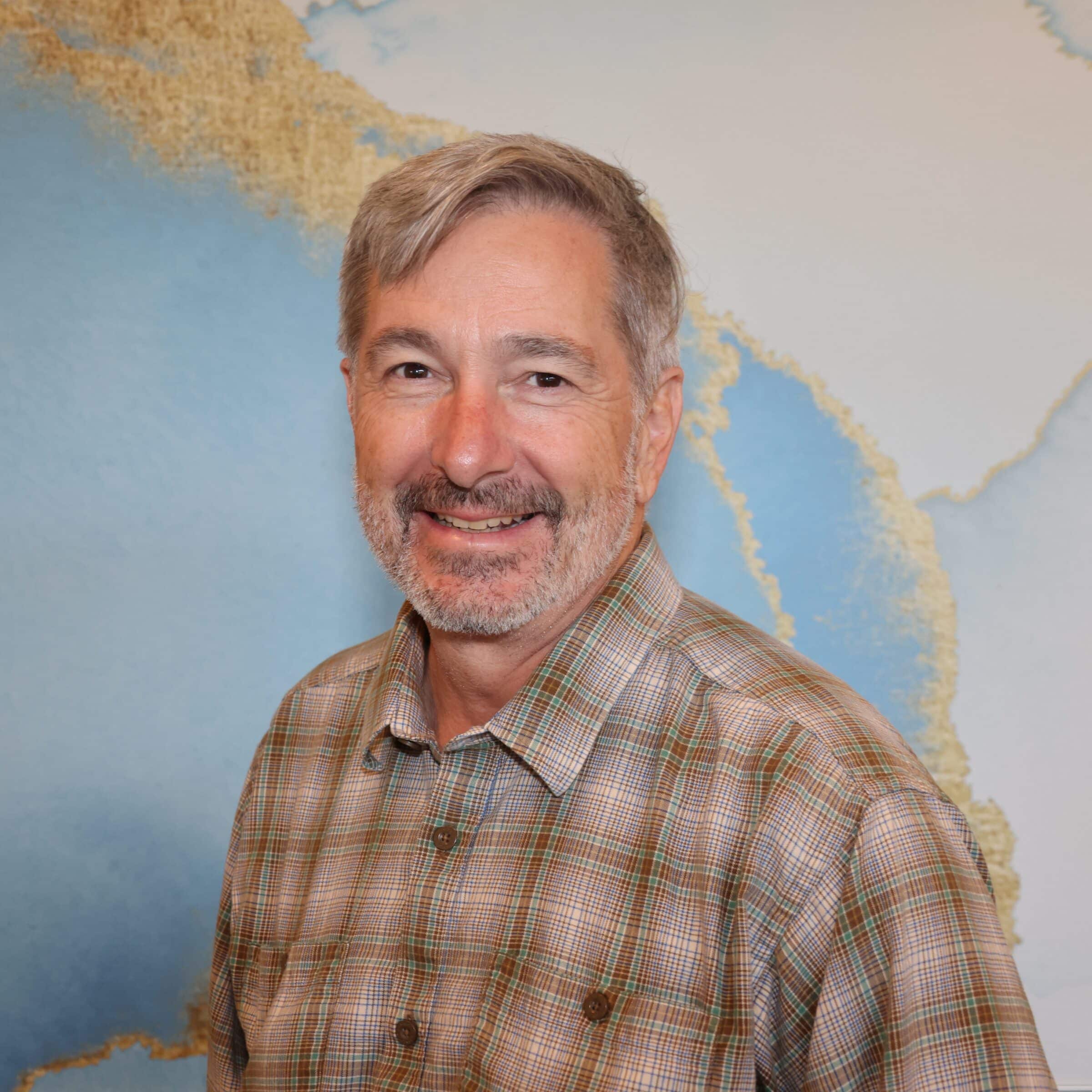 A middle-aged man with gray hair and a beard, wearing a plaid shirt, smiles at the camera. A large map with blue and tan colors serves as the background.