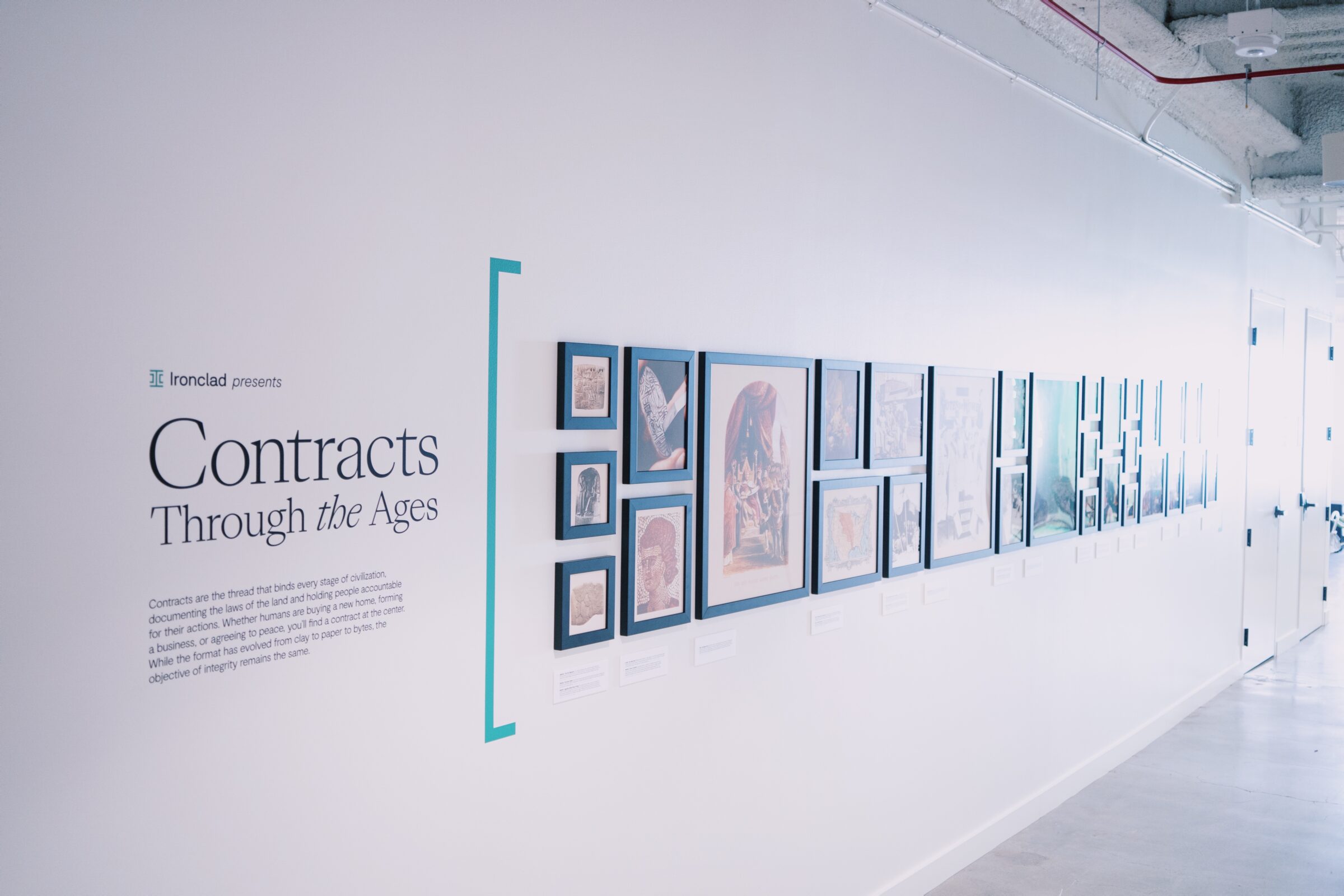 A white hallway features framed documents and images displayed in a row on the wall, with a sign reading Contracts Through the Ages introducing an exhibit about the history of contracts.
