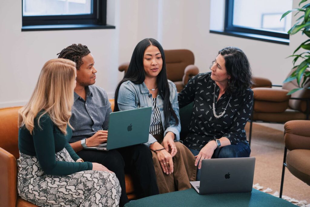 Four people sit together on a couch having a discussion, each with a laptop open in a modern office setting. They appear engaged and are casually dressed.