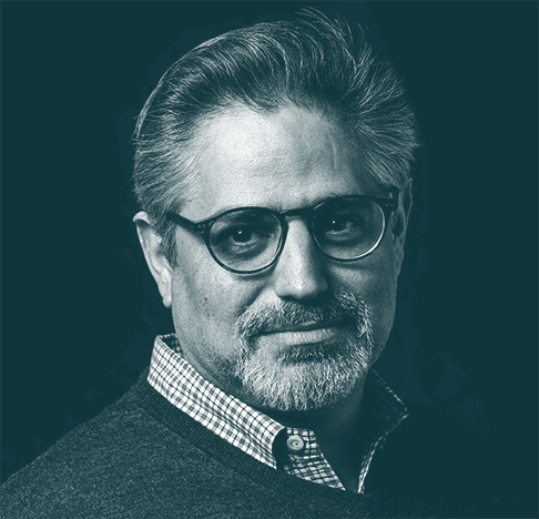 A middle-aged man with gray hair, a beard, and glasses, wearing a checkered shirt and a dark sweater, looks into the camera against a dark background.