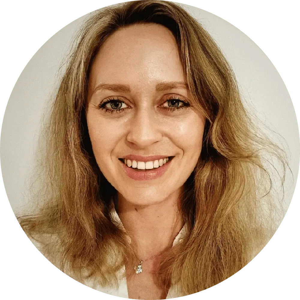 A woman with long, wavy blonde hair smiles at the camera in an outlaw alternative style. She wears a light-colored top and a delicate necklace with a small pendant, set against a plain white background within a circular frame.