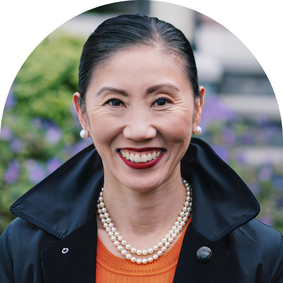 A woman with dark hair pulled back, wearing a black coat, orange top, pearl necklace, and pearl earrings, smiles outdoors with blurred greenery and purple flowers in the background.
