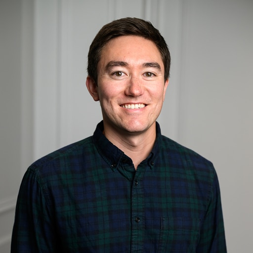 A person with short dark hair, wearing a dark green and blue plaid button-up shirt, smiles while standing indoors in front of a plain light-colored wall.