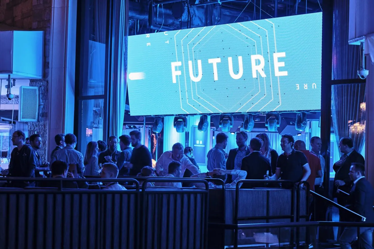 A group of people socializes in a dimly lit venue with blue lighting. A large digital screen above them displays the word FUTURE in bold letters with geometric designs.