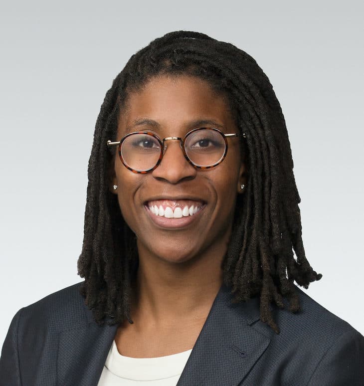 A person with medium-length dreadlocks, wearing round glasses, a navy blazer, and a white top, smiling in front of a plain light gray background.