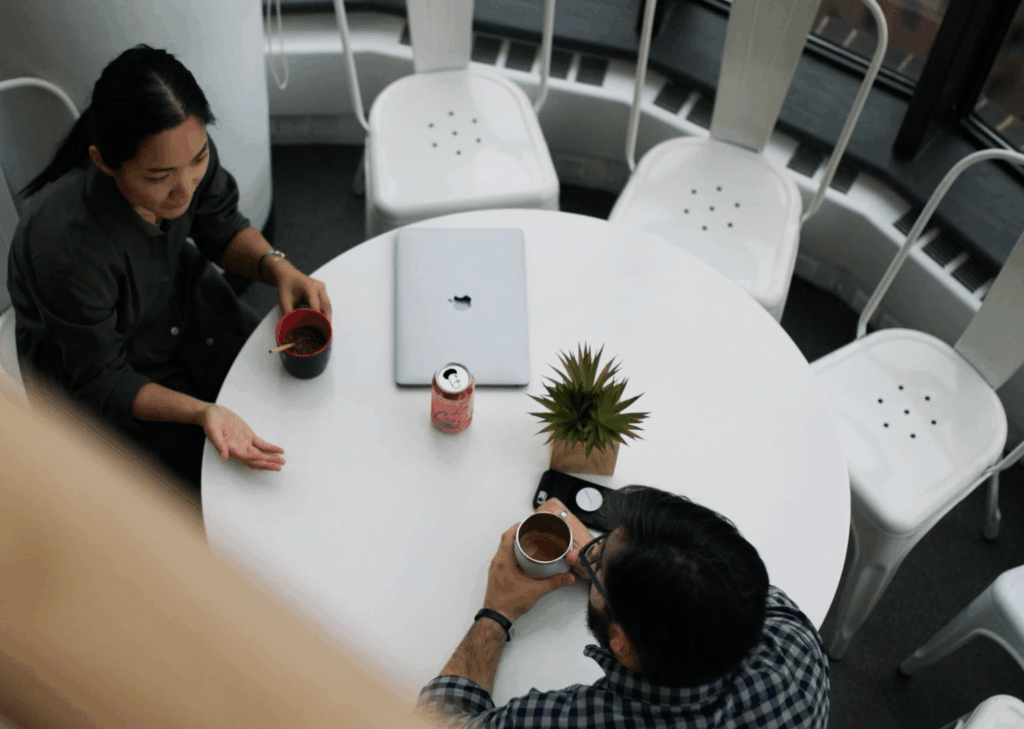 Two people sit at a round white table with cups of coffee, a soda can, a closed laptop, and a small potted plant. White chairs surround the table, and the scene is lit by natural light from large windows.