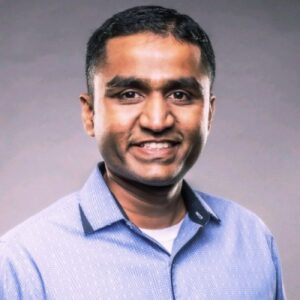 A man with short black hair smiles at the camera. He is wearing a light blue button-up shirt over a white undershirt. The background is plain and neutral-colored.