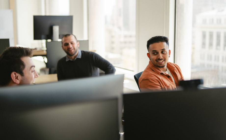 Three men in a modern office sit at desks with computer monitors, engaged in conversation about using AI for career growth and smiling. Large windows let in natural light, with city buildings visible outside.