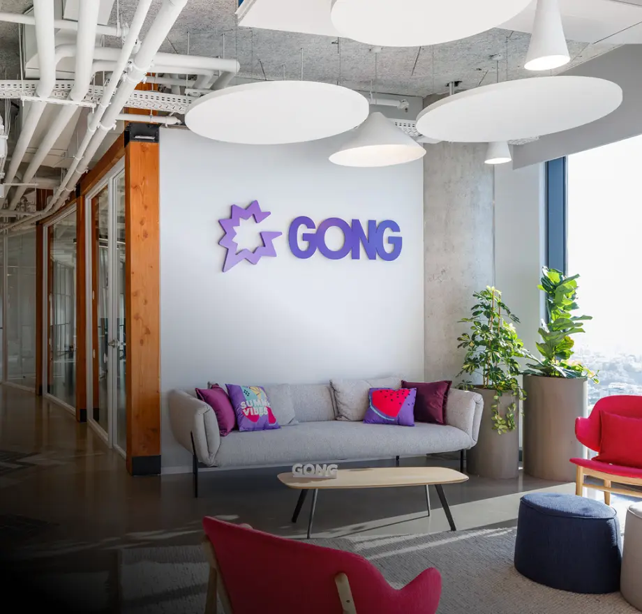 Modern office lobby with a gray couch, colorful pillows, a red chair, potted plants, and a coffee table. The wall displays a large purple GONG logo. Large windows let in natural light.