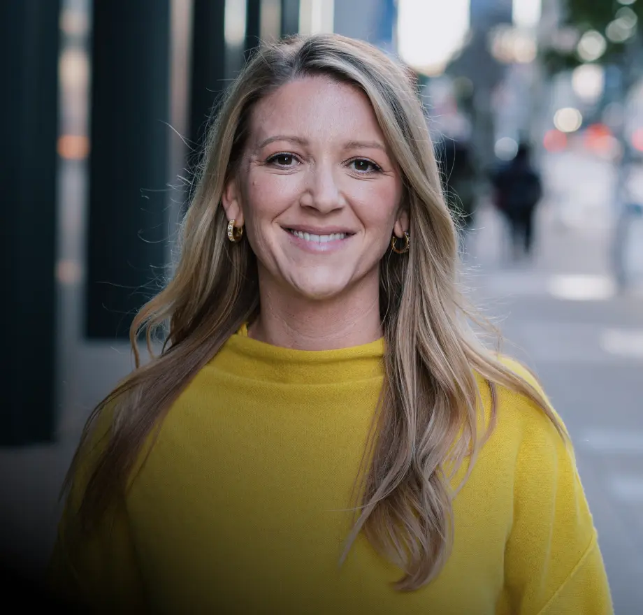 A woman with long blonde hair, wearing a yellow sweater and hoop earrings, smiles while standing outside on a city sidewalk.
