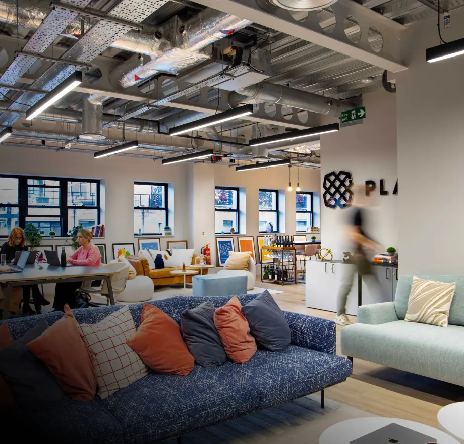 Modern open-plan office with sofas, desks, and people working on laptops. Exposed ceiling pipes and large windows let in natural light. Blurred figure walks past, creating a dynamic atmosphere. Pillows decorate the seating area.