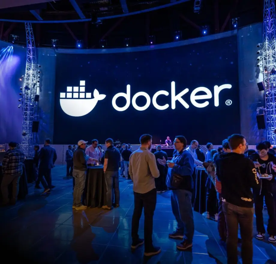 A group of people network at an indoor tech event with a large illuminated Docker logo displayed on a screen in the background.