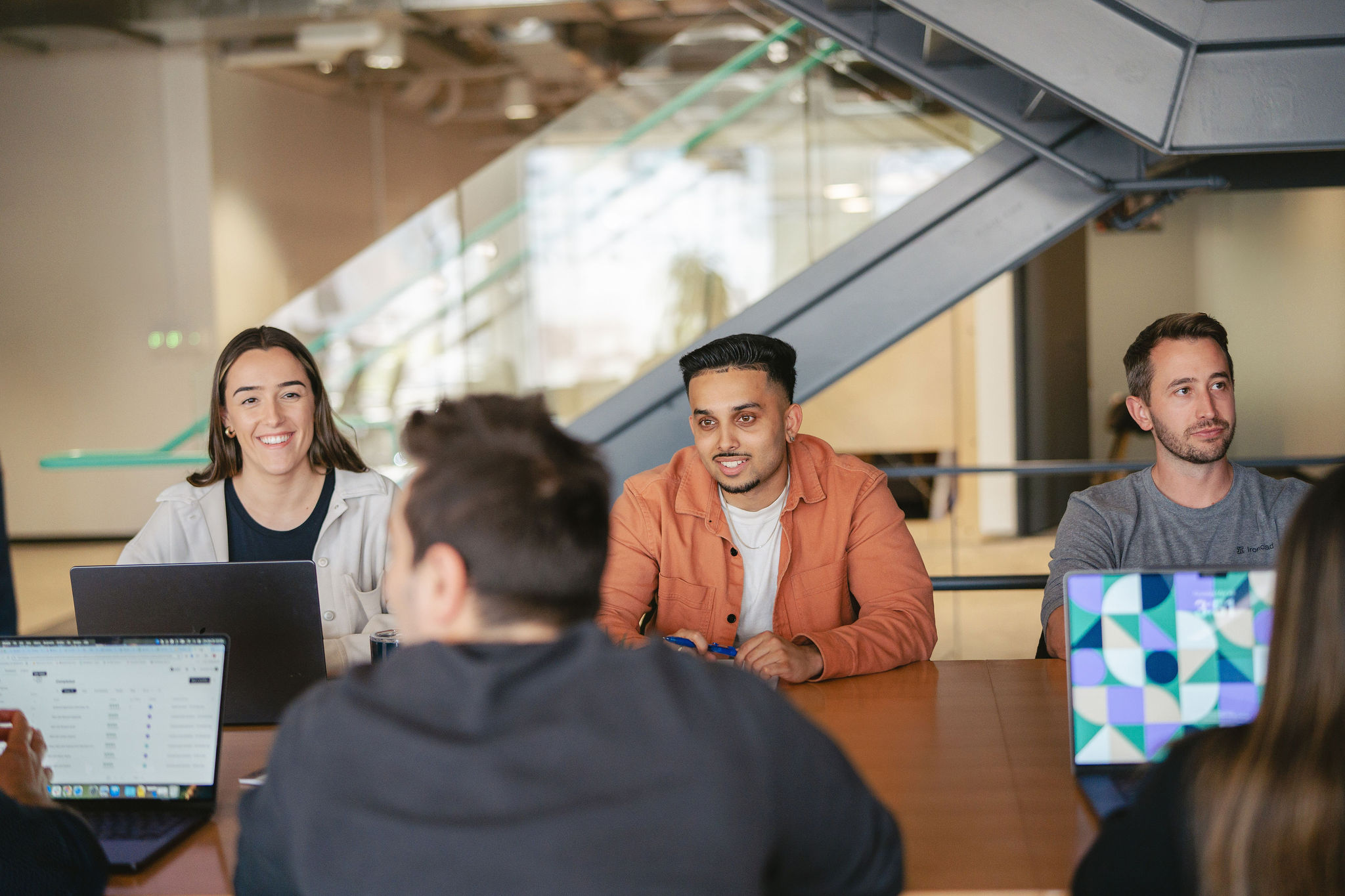 Four people sit around a wooden table with laptops in an office, engaged in discussion. Two women and two men are visible, with a staircase and glass wall in the background.
