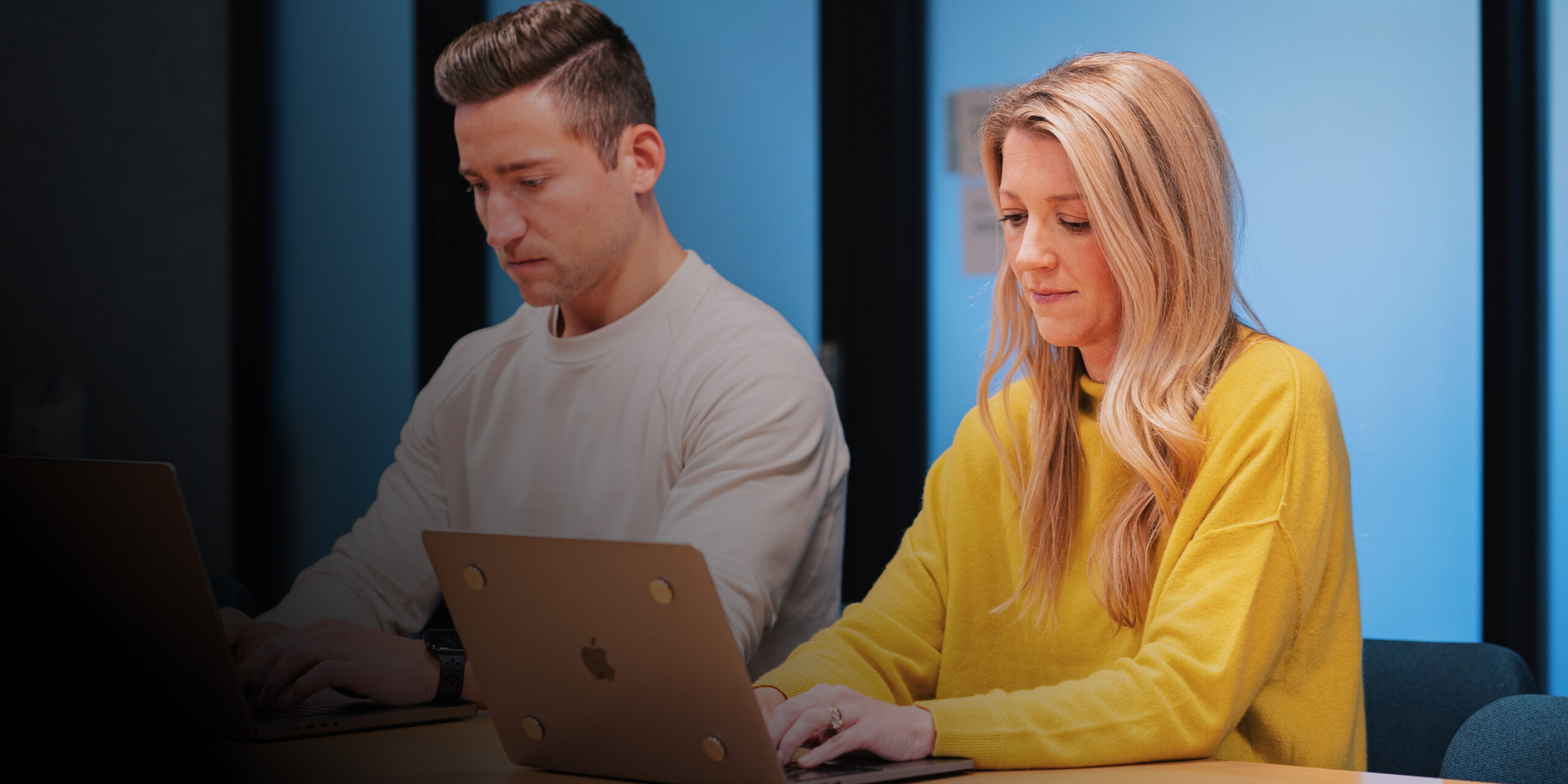 Two people sit side by side at a table, working on laptops like ironclad partners. The man on the left wears a white shirt, while the woman in a yellow sweater sits on the right. Both focus intently in a modern office setting.