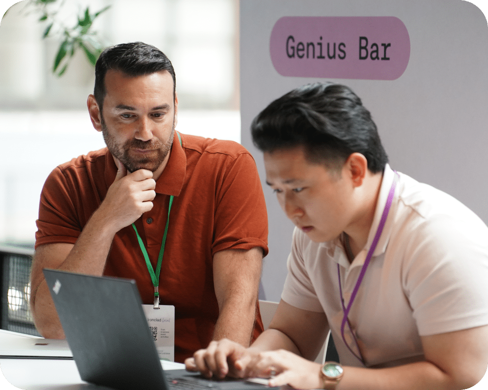 Two men sit at a table labeled Genius Bar. One man in a rust-colored shirt listens attentively while the other, in a white shirt, focuses on typing on a laptop. Both wear lanyards and appear engaged in discussion.