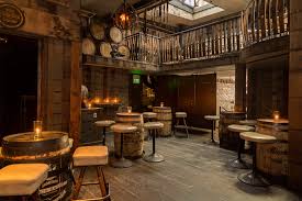 A rustic bar interior with wooden barrels used as tables, surrounded by stools. Warm candlelight creates a cozy atmosphere, and a wooden balcony overlooks the main seating area.