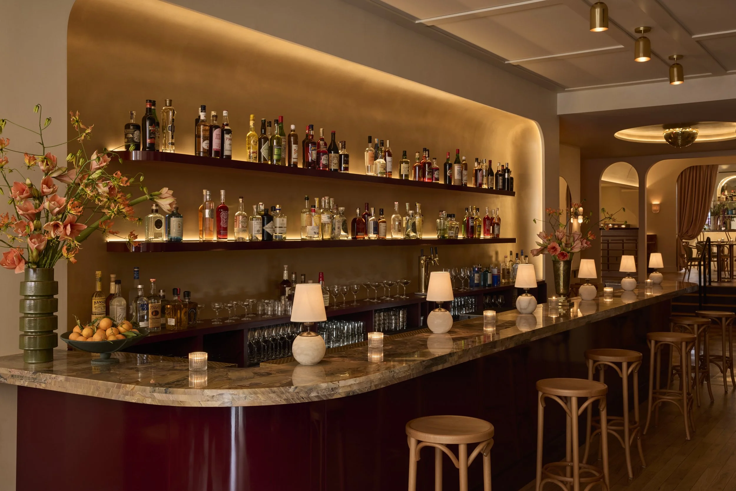 A stylish bar with a marble countertop, wooden stools, small lamps, candles, and floral arrangements. Shelves behind the bar display various bottles of alcohol against a softly-lit golden wall. The atmosphere is warm and inviting.