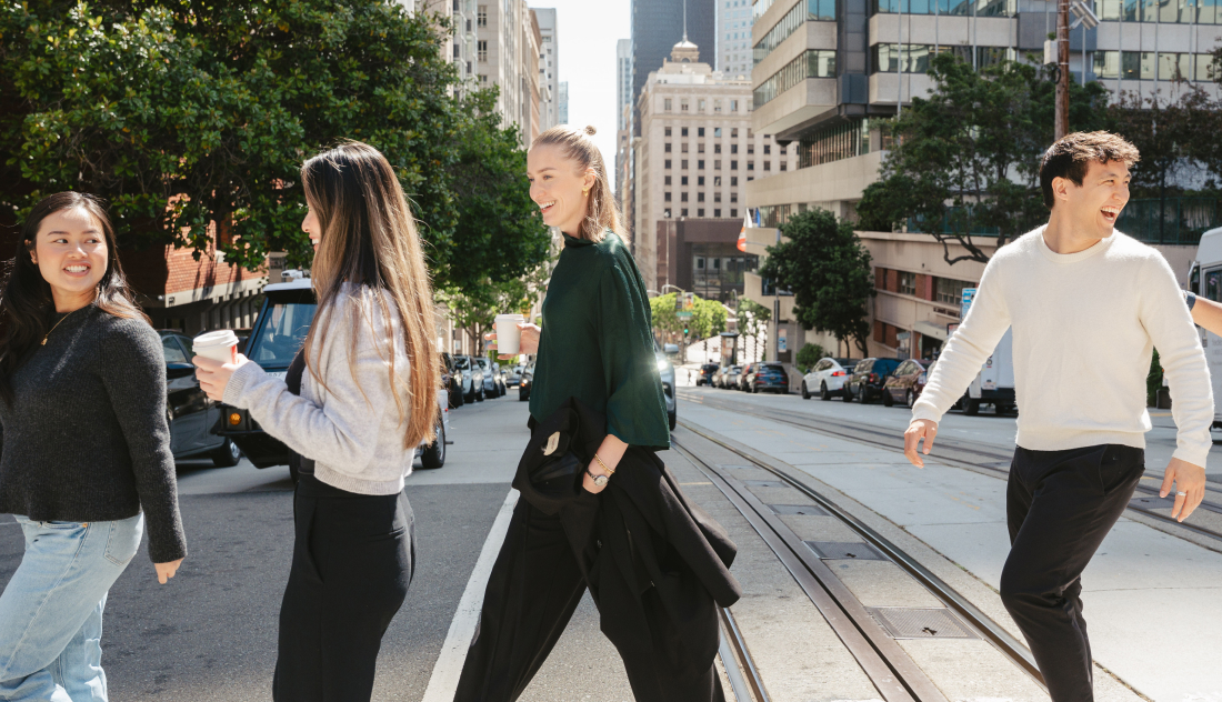 Four young adults cross a city street on a sunny day, smiling and holding coffee cups, with tall buildings and trees lining the background.