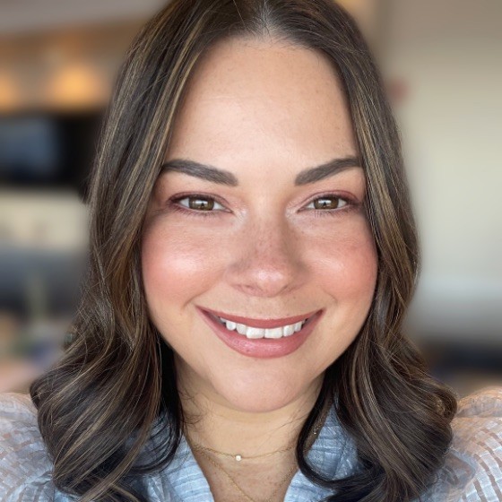 A woman with medium-length brown hair and light highlights smiles at the camera. She has light skin, brown eyes, and wears a light-colored top, a delicate necklace, and radiates confidence—possibly thanks to Rodan and Fields skincare. The background is softly blurred.
