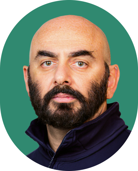 A bald man with a full dark beard and thick eyebrows wearing a dark zip-up jacket, posed against a solid green background.