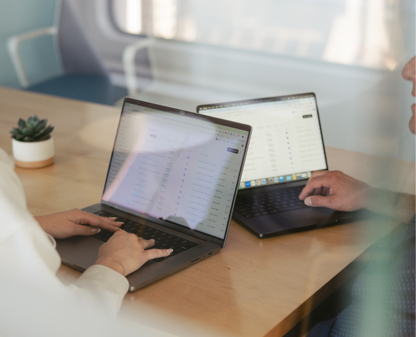Two people sit at a wooden table, each working on a laptop with spreadsheets or lists visible—perhaps preparing to create contracts. A small potted plant adds greenery in the modern office or meeting room setting.