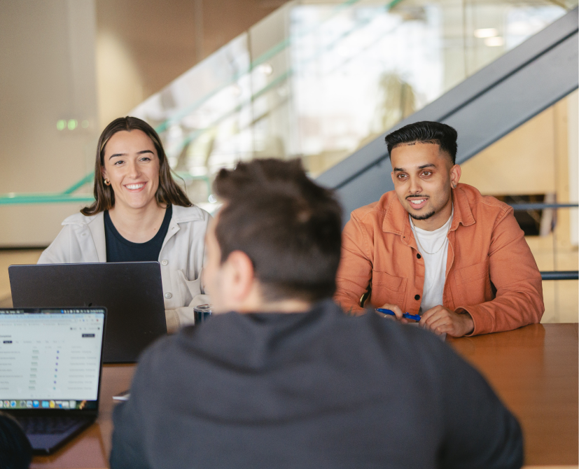 Three young adults sit at a table in an office, two facing the camera and smiling with laptops open, as they collaborate to create contracts. A staircase and glass railing are visible in the background.