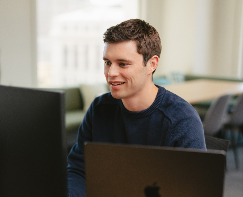 A young man in a blue sweater sits at a desk, smiling while working on a computer and creating contracts with another laptop in front of him in a bright office setting.