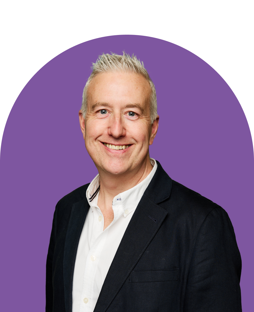 A smiling man with short gray hair, wearing a dark blazer over a white shirt, poses in front of a purple background, ready to create contracts with confidence and professionalism.