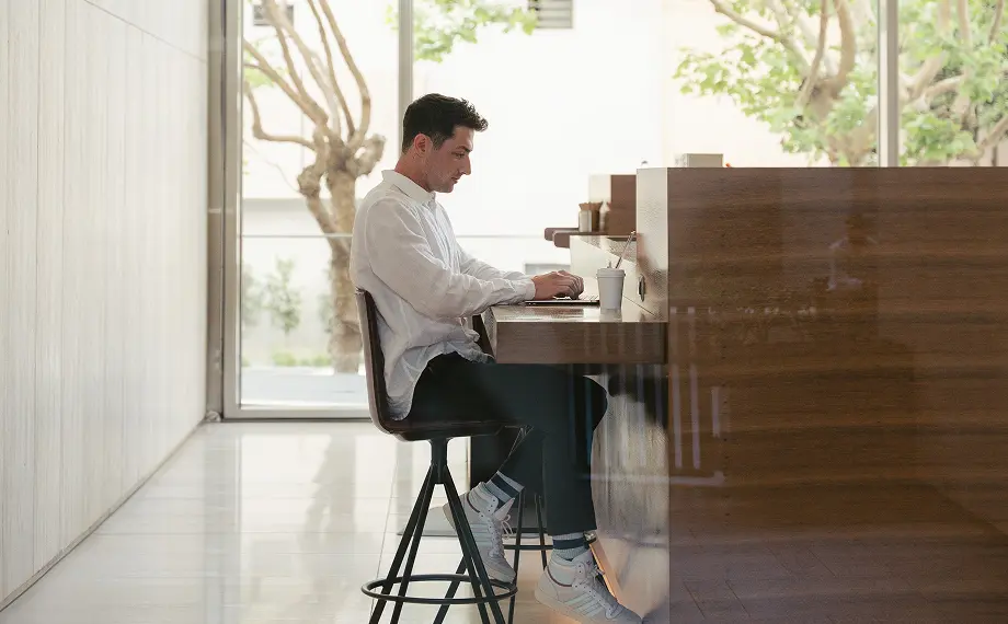 A person in a white shirt sits on a high stool at a wooden counter, typing on a laptop in a bright, modern space with large windows and trees outside, perhaps researching contract management best practices.