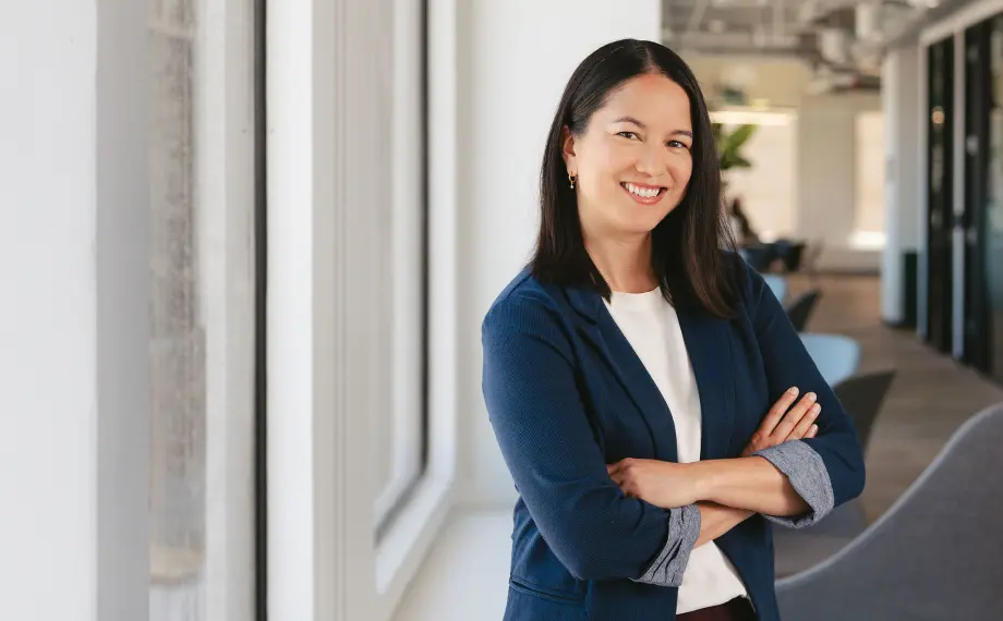 Woman with long dark hair, wearing a blue blazer and white shirt, stands indoors with arms crossed, smiling at the camera—portraying the confidence of someone who knows how to become a legal operations manager. Bright windows and a modern office setting are visible in the background.
