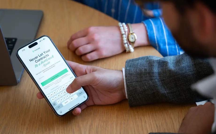 A person in a suit types on a smartphone displaying an influencer agreement within a contract management app, while another person with a watch and bracelets rests their hand on a table nearby.