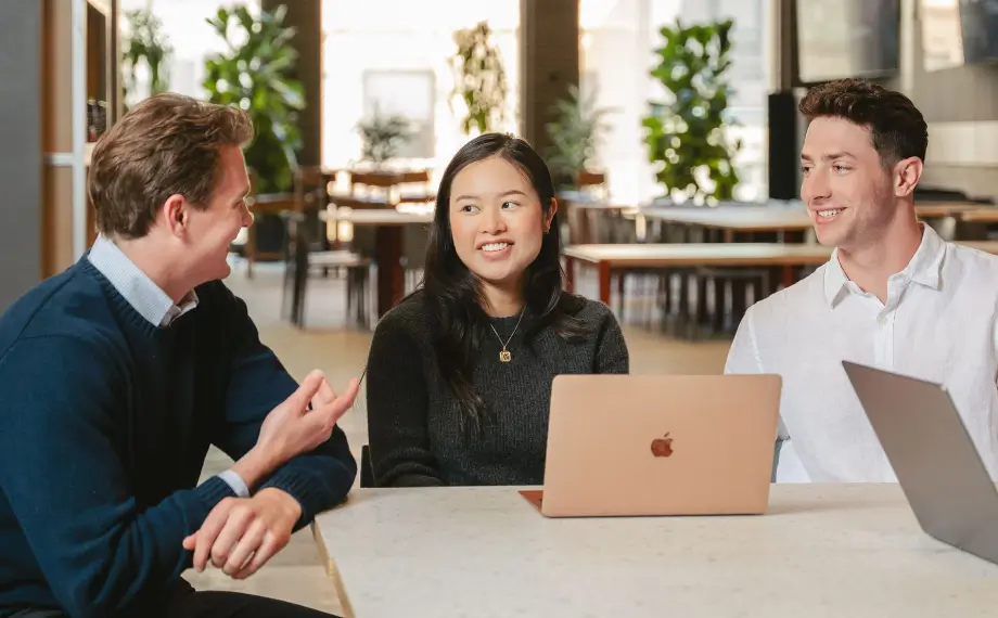 Three people sit at a table with laptops, smiling and talking in a modern, bright office space with plants and large windows in the background. Their discussion highlights how procurement contract management is not just a legal responsibility but also an opportunity to foster collaboration and efficiency within their organization.