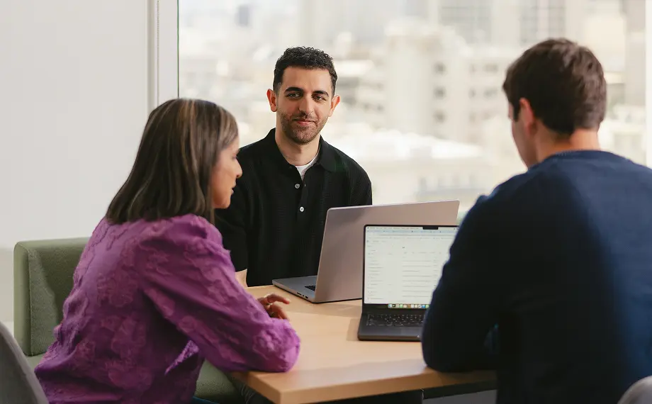 Three people sit around a table in an office with laptops open, likely discussing a content licensing agreement. Two are facing away, while one man faces the camera. A cityscape is visible through the window behind them.