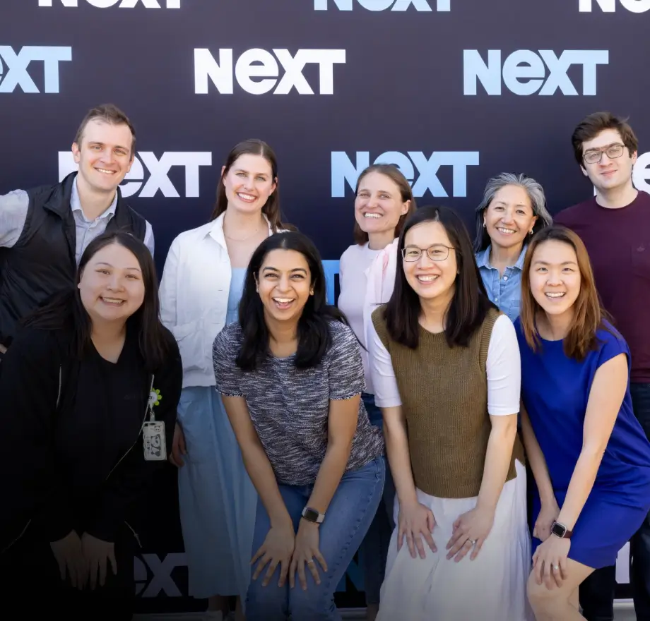 A group of nine people smiling and posing together in front of a backdrop with the word NEXT repeated on it.