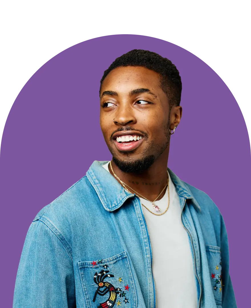 A smiling man in a denim jacket and white shirt looks to the side, radiating confidence as someone ready to create contracts. He has short hair, a trimmed beard, and wears a gold necklace. The background is white with a purple half-circle behind him.