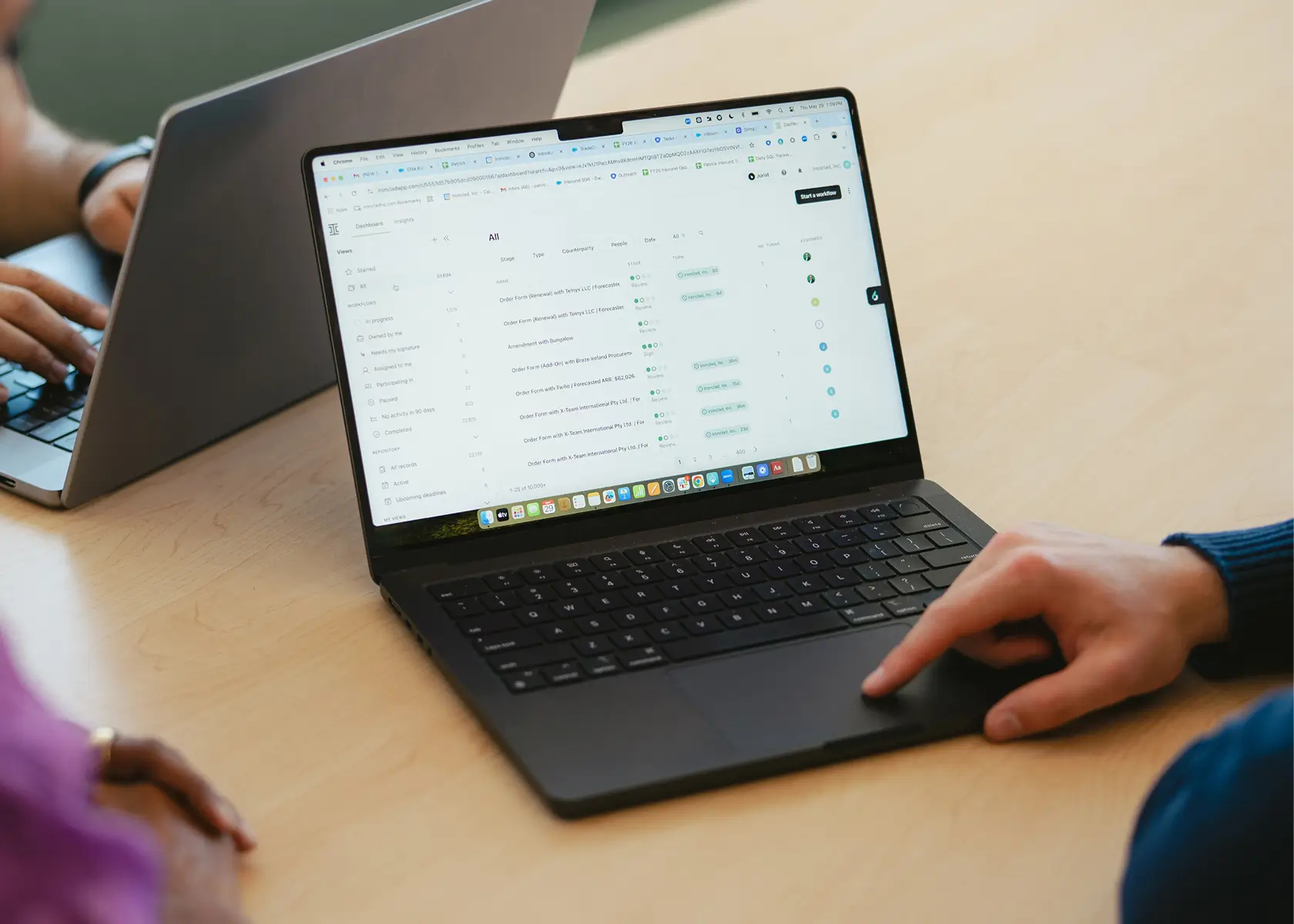 Two people work at a table with laptops. One person points to a screen displaying a list of emails or messages, while the other person types on their own laptop in the background.