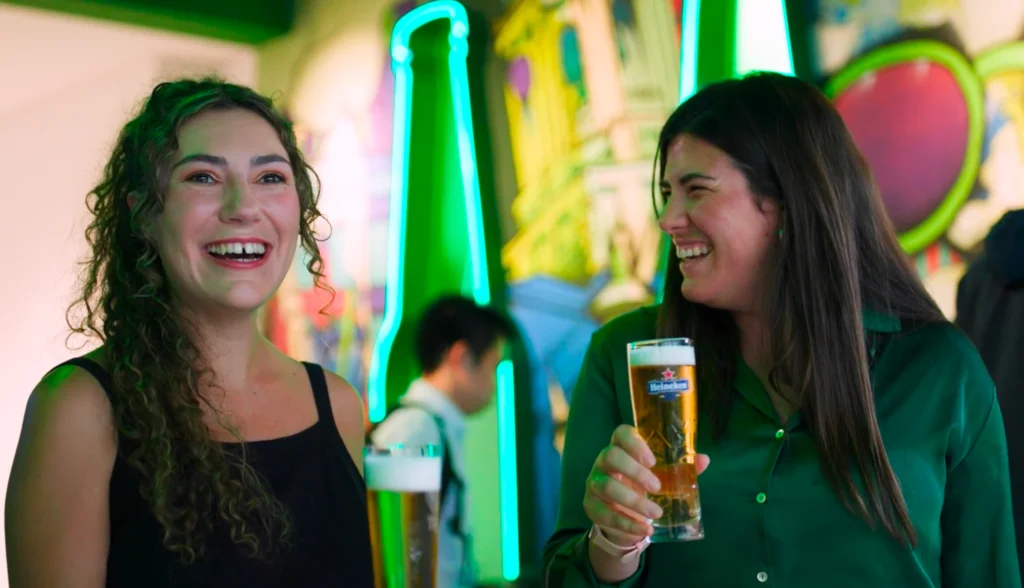Two women smiling and holding glasses of beer in a lively, colorful bar with neon green lights and ironclad wall art in the background.