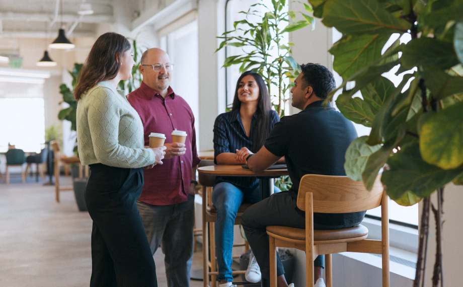 Four people in a modern office space talk and smile near a table with drinks. Two are seated, two are standing, holding coffee cups, surrounded by large green plants and natural light from big windows.