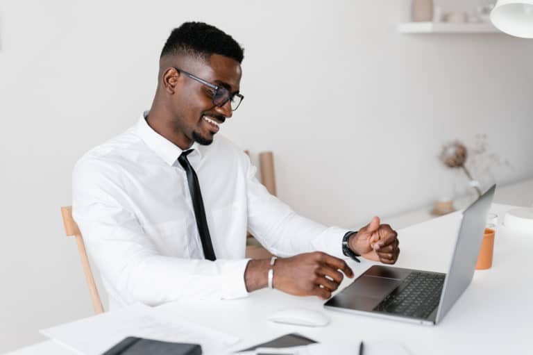 man in glasses smiling and browsing laptop while sitting at table and working in contemporary workplace