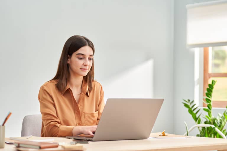 Confident young woman in casual clothes sitting at table and browsing laptop while working on remote project in light home office | How to create a clickwrap agreement